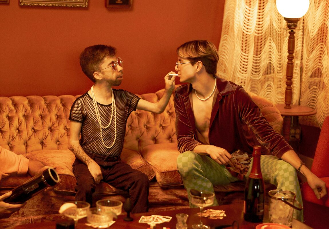 Deux hommes assis dans un salon à l'ambiance 70s. L'un deux met une cigarette dans la bouche de l'autre.