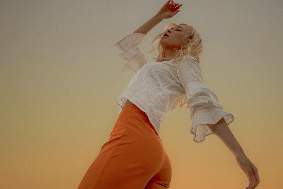 personne blonde portant une chemise blanche et des pantalons orange et qui semble danser.
